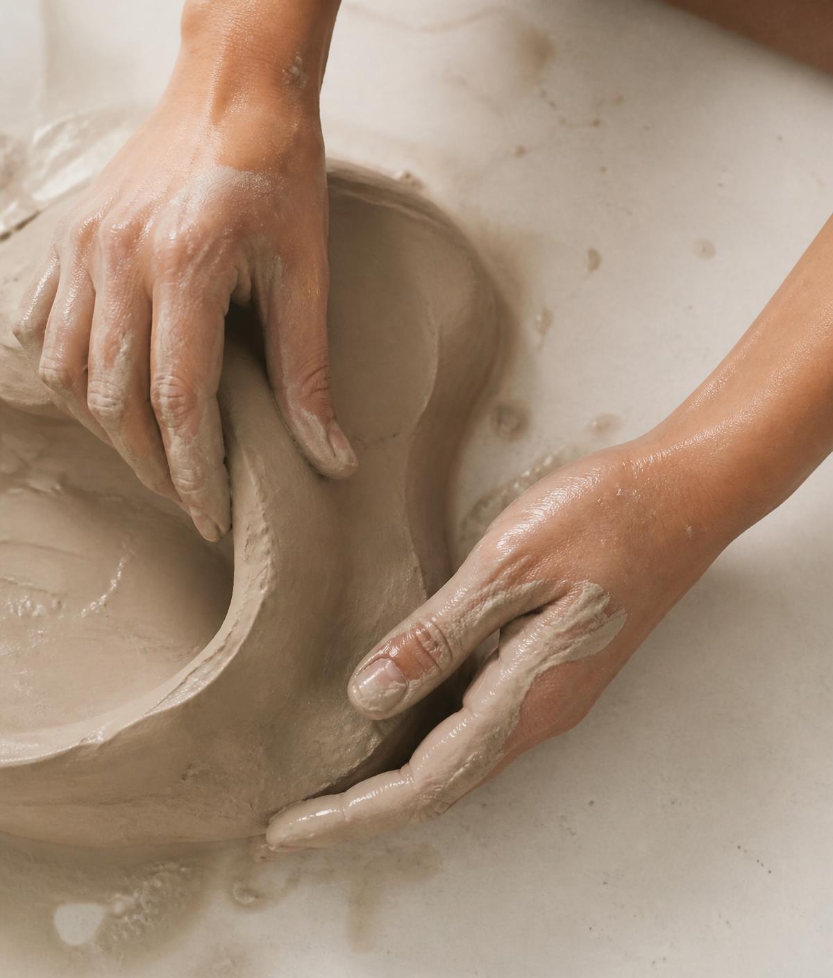 Hands shaping wet clay on a pale surface, the material taking an individual form